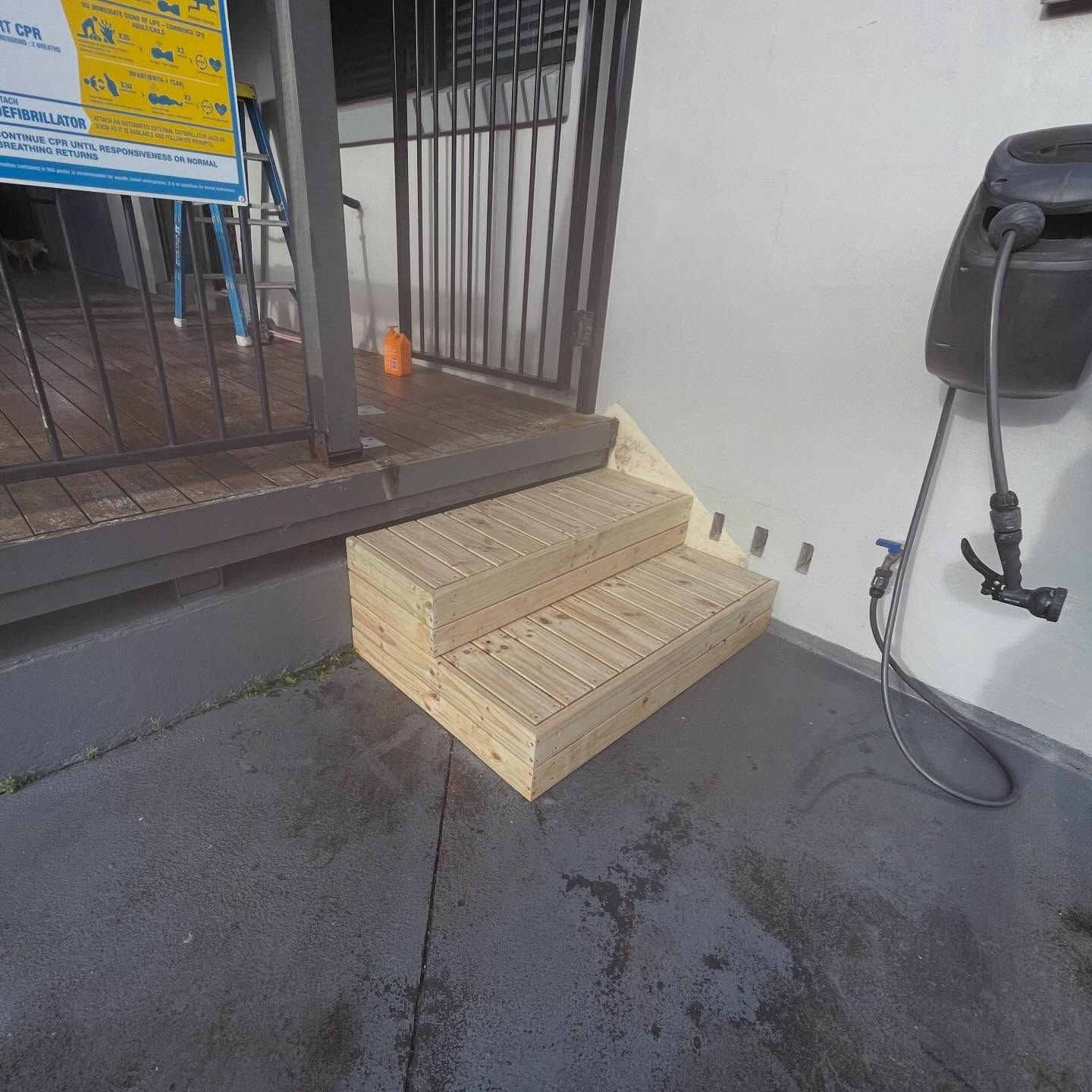New wooden two-step staircase on a grey concrete patio next to a wall with an EV charging station.