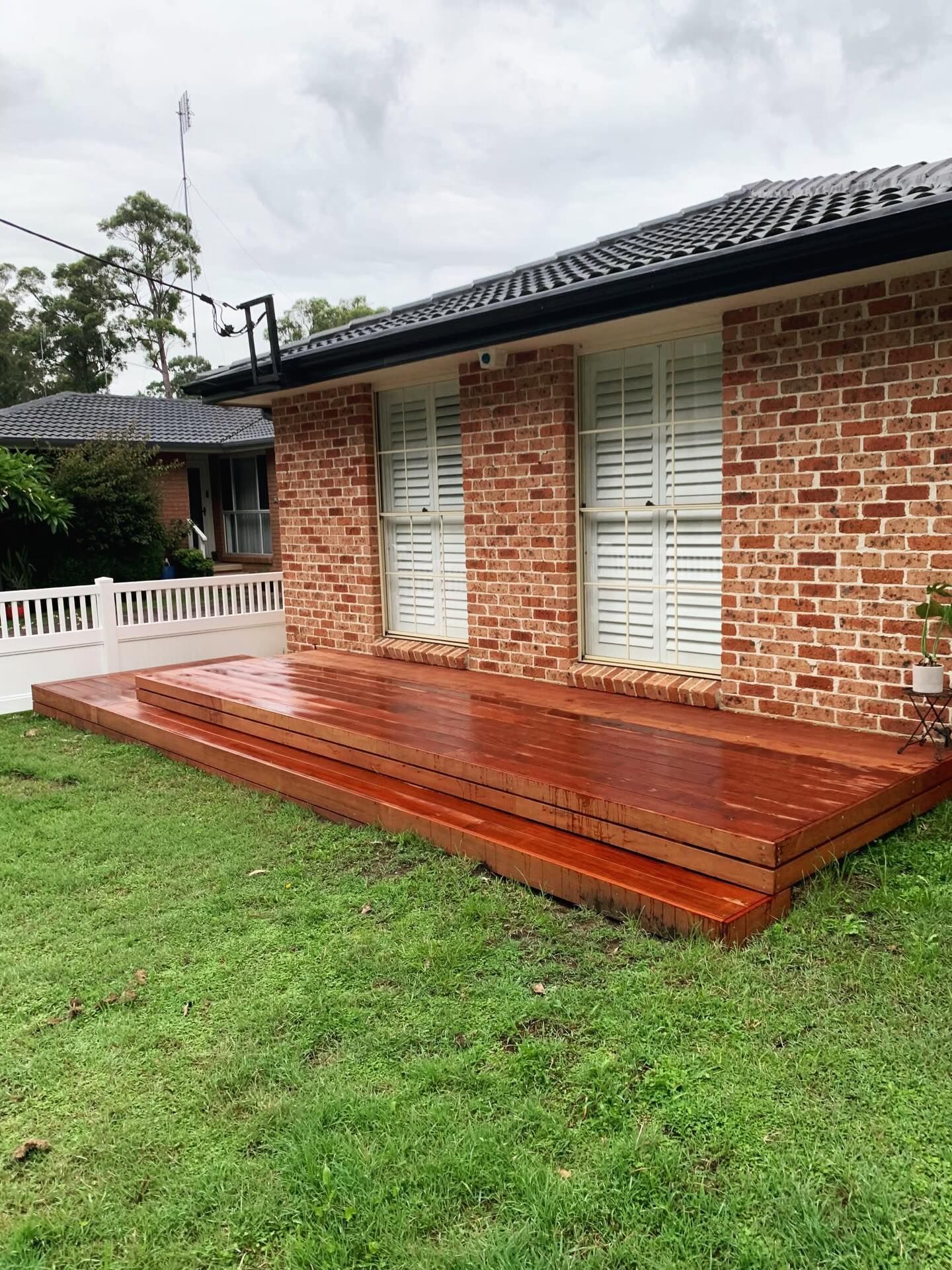 A newly stained reddish-brown wooden deck attached to the side of a brick house with white window shutters on a lawn.