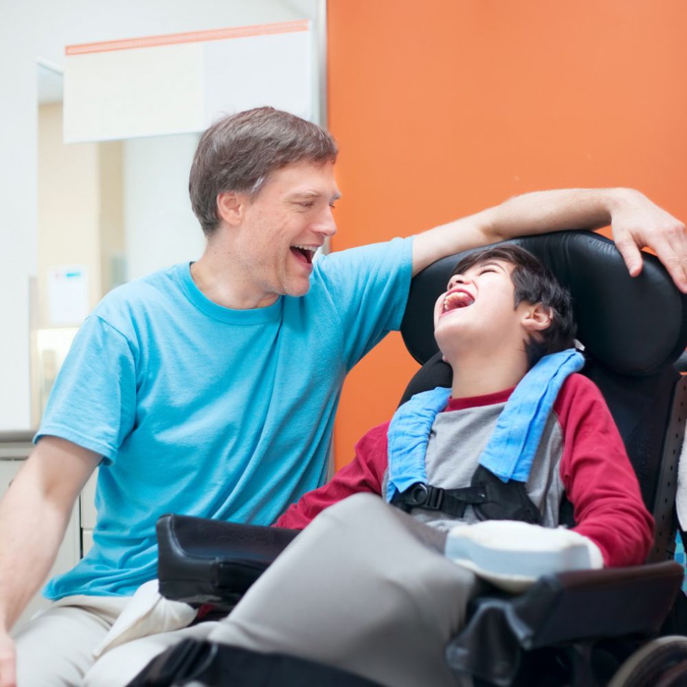 A man in a blue shirt is laughing with a boy in a wheelchair