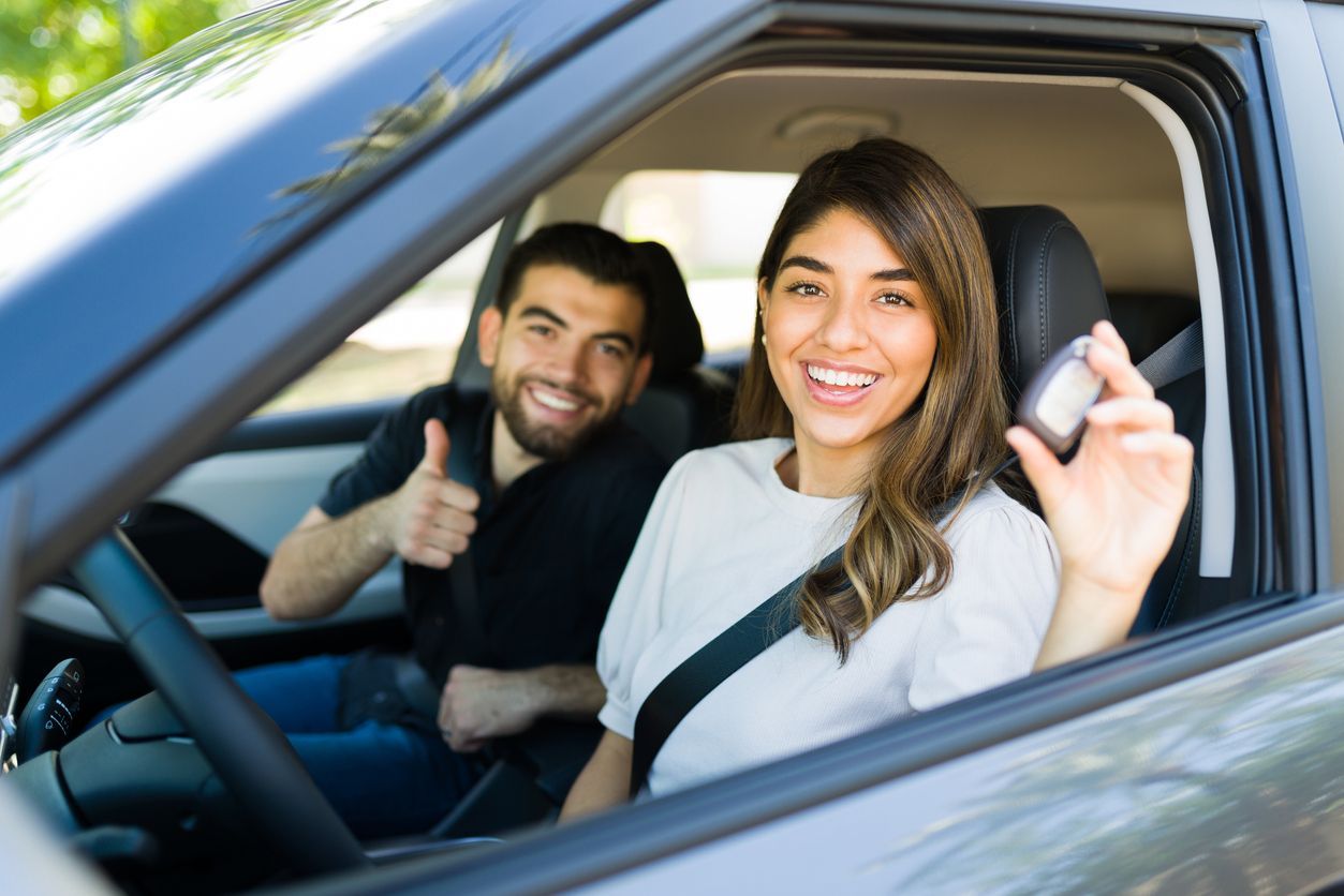 Una mujer en un coche con las llaves, sonriendo. Un hombre al fondo levanta el pulgar, ambos felices.