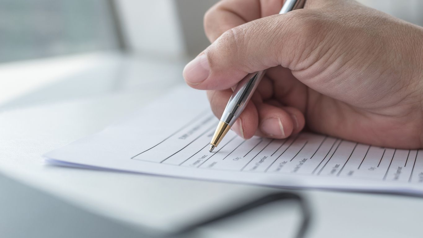 Mano de una persona sosteniendo un bolígrafo, escribiendo en un formulario con líneas, sobre una superficie blanca.