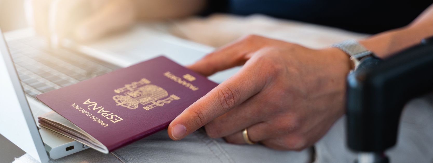 Una mano sostiene un pasaporte español sobre un portátil. Lleva reloj y anillo de bodas.
