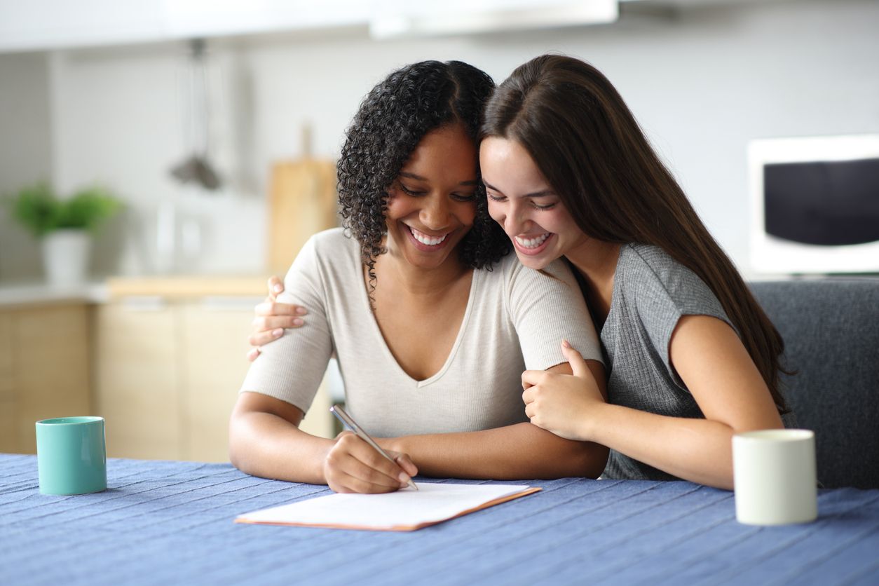 Dos mujeres sonrientes, una escribiendo en una mesa mientras la otra la abraza.