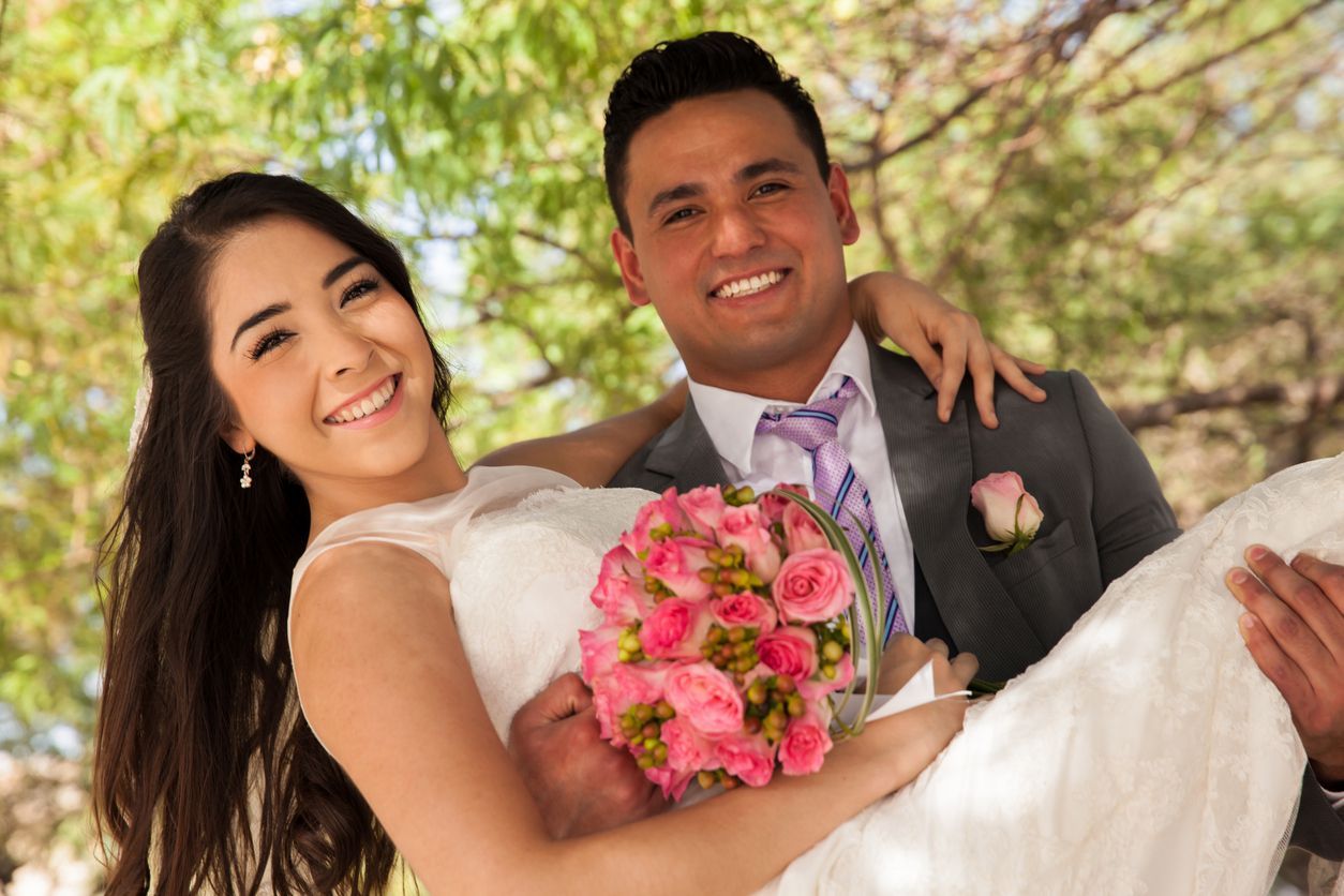 El novio carga a la novia, ambos sonriendo. La novia sostiene flores rosas, con un vestido blanco. Al aire libre, soleado.