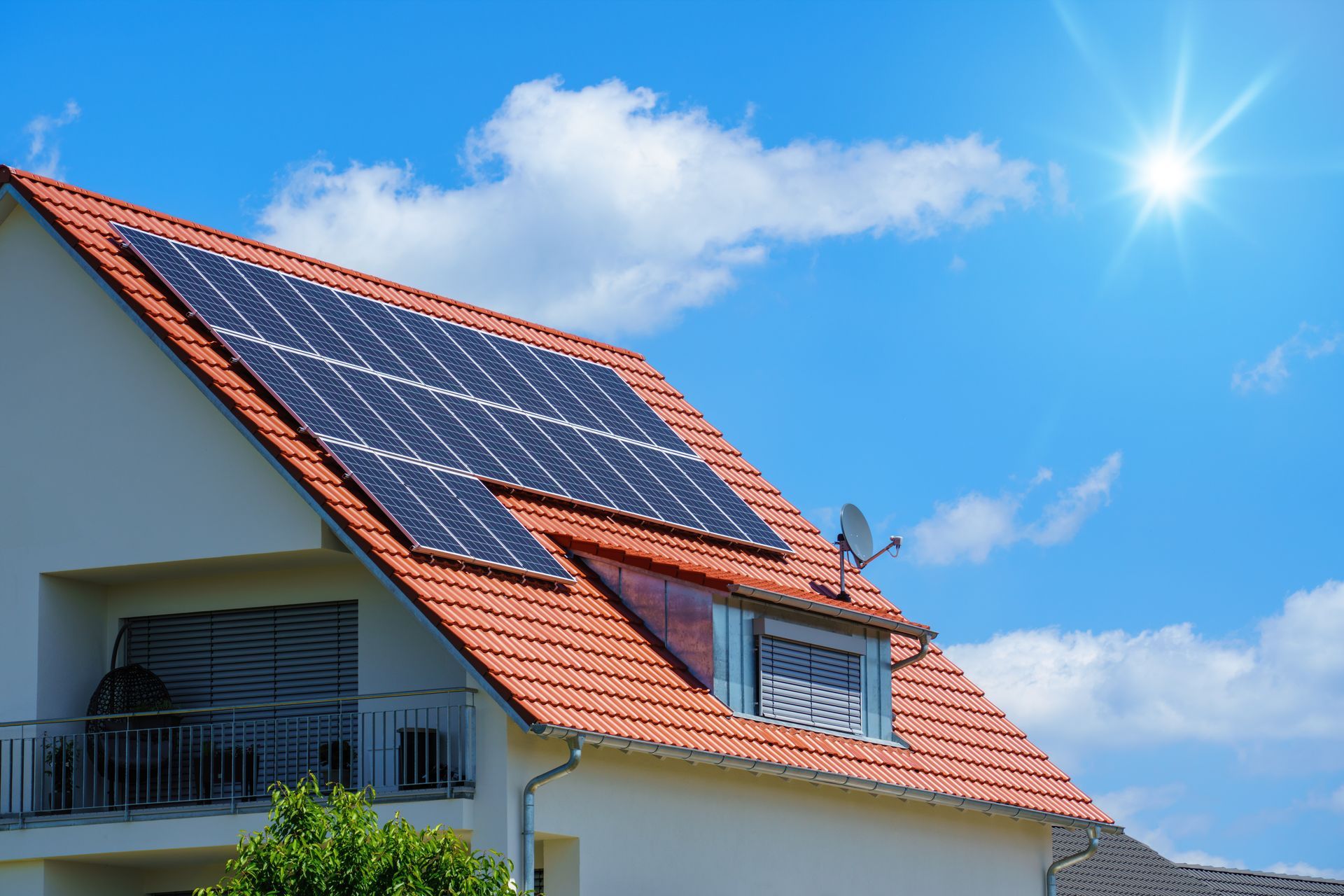 Solar panels on a red-tiled roof of a house, under a bright sun and blue sky.