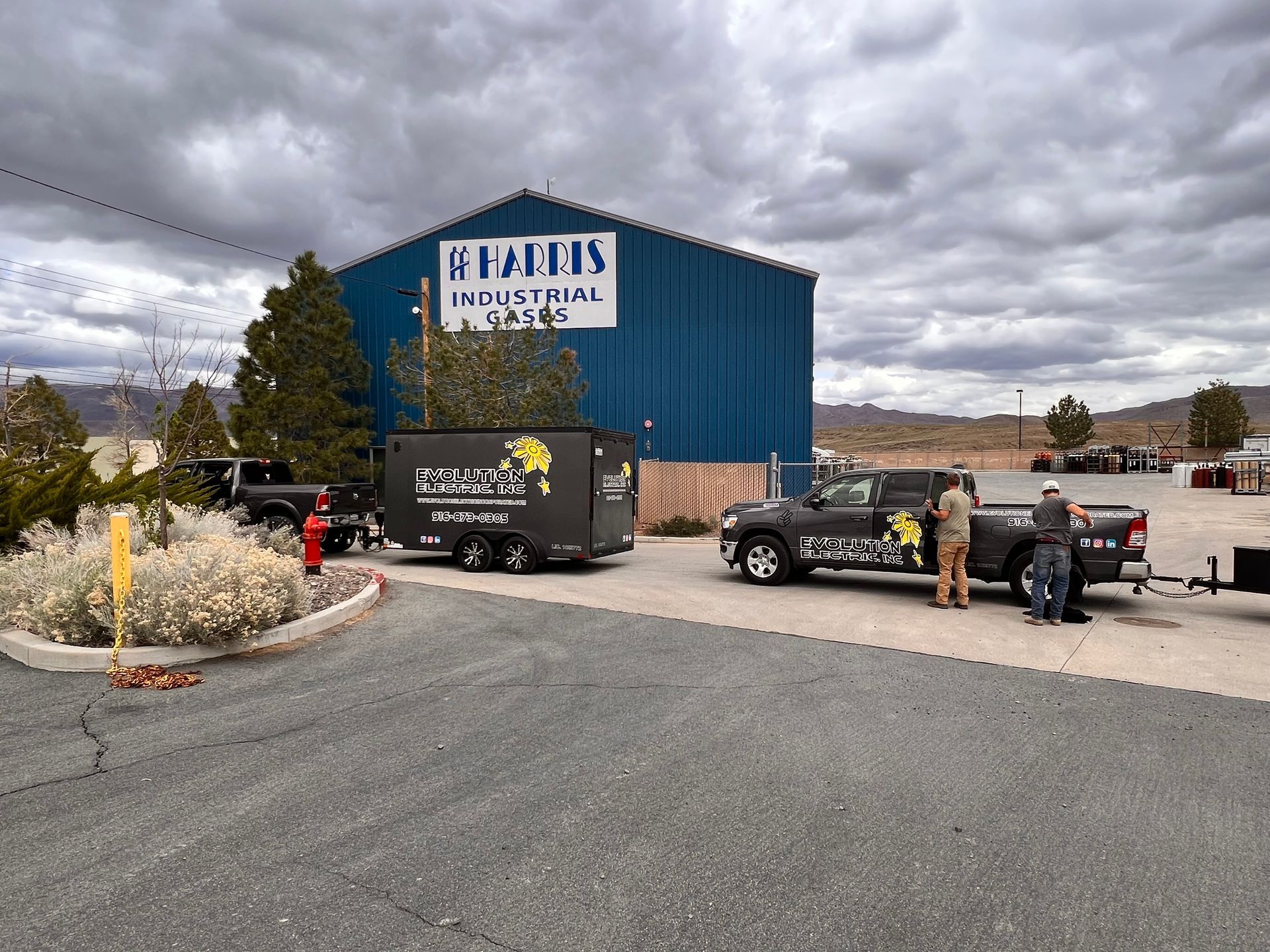 Two trucks with trailers outside a blue industrial building under a cloudy sky.