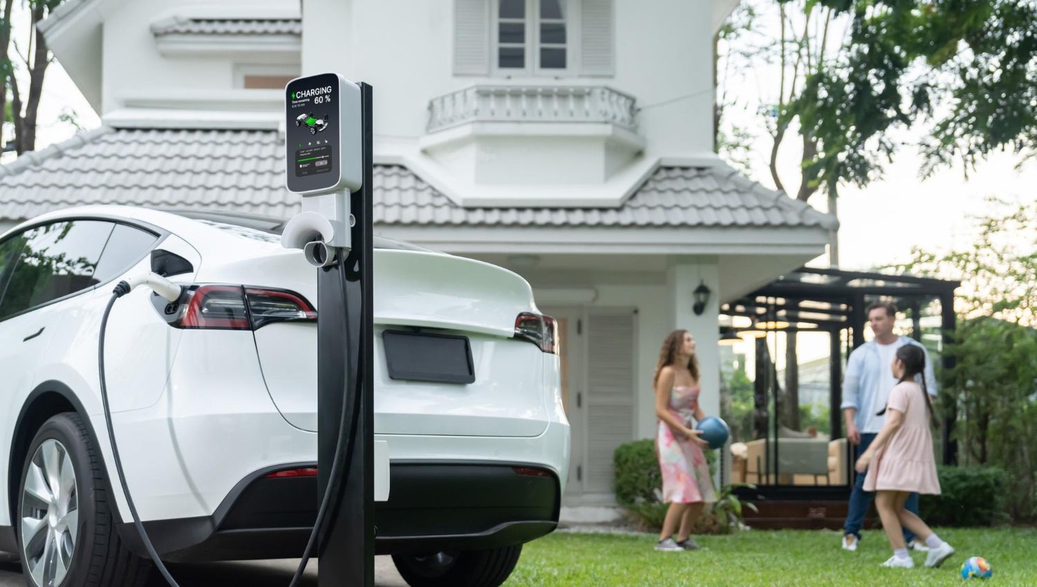 A white electric car is being charged at a charging station in front of a house.