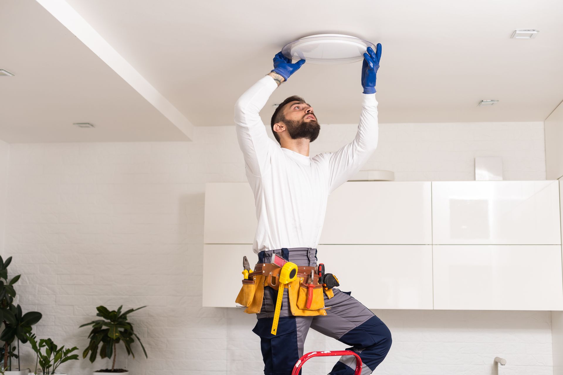 Person in work clothes installing a ceiling light fixture on a ladder indoors.