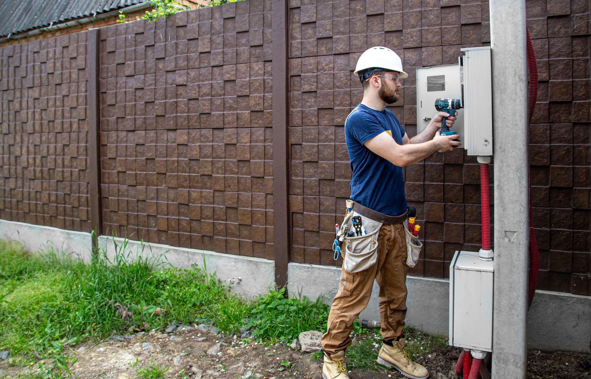 Electrician in a hard hat working on an electrical panel attached to a concrete post next to a decorative brown fence.