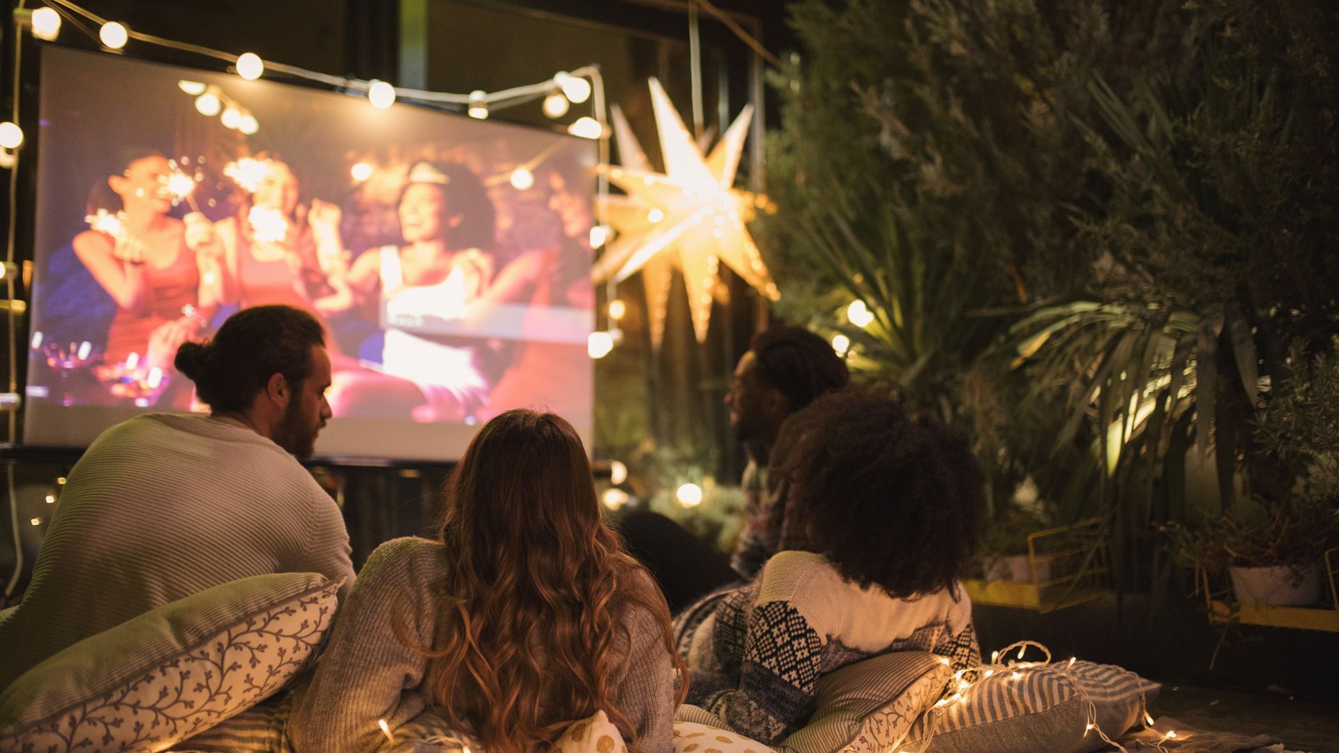 People watching a movie outdoors on a screen at night, lit by string lights and a star.