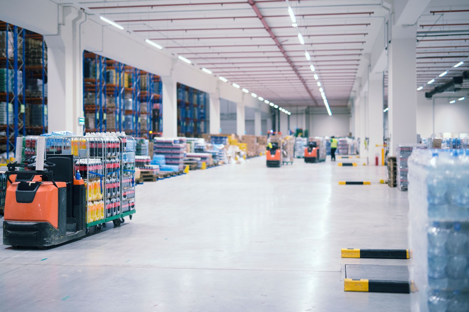 Warehouse interior with forklift carrying goods, rows of shelves stacked with products.