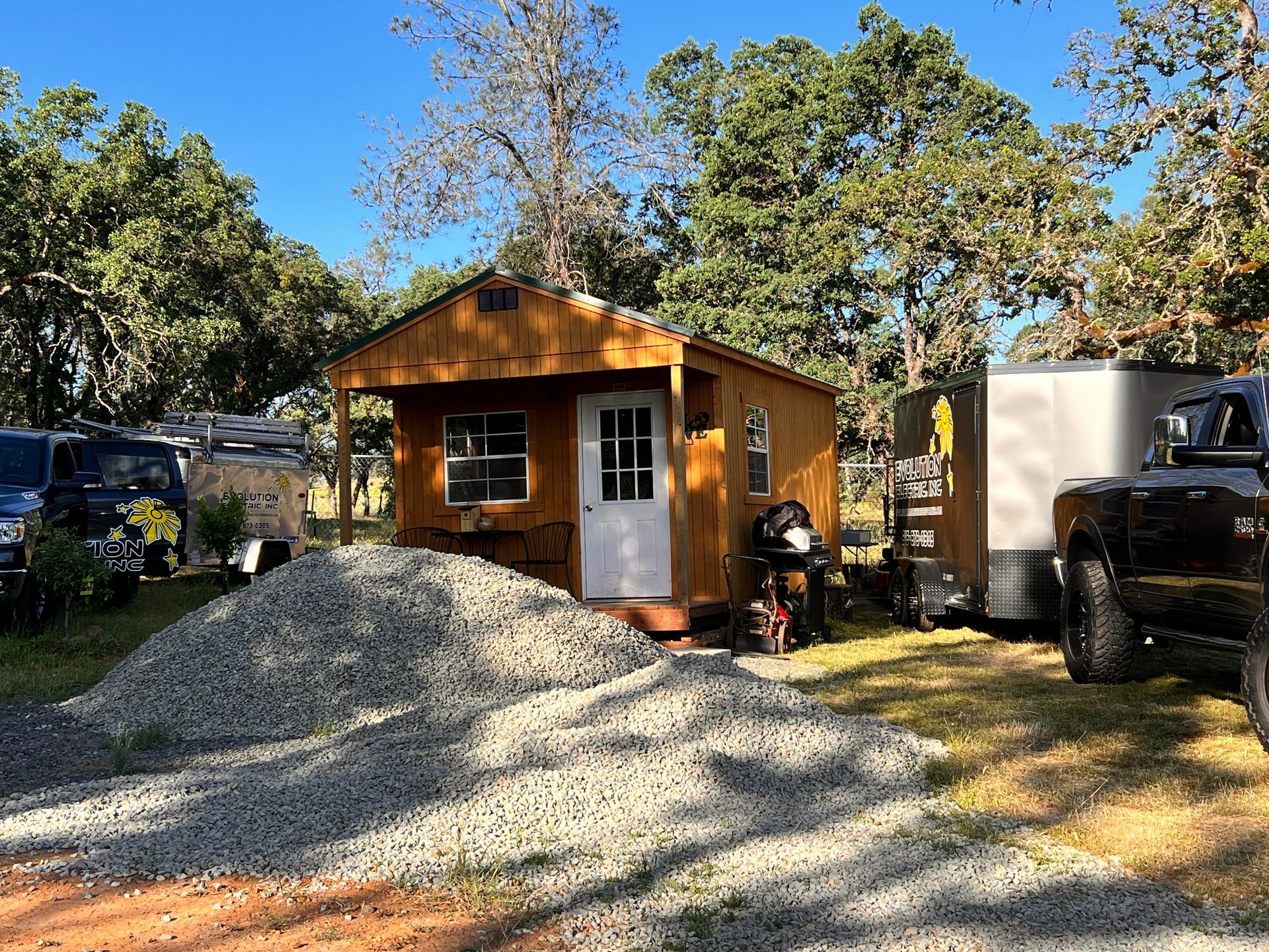 A small, wooden cabin with a gravel pile in front. Trees and trailers surround the structure on a sunny day.