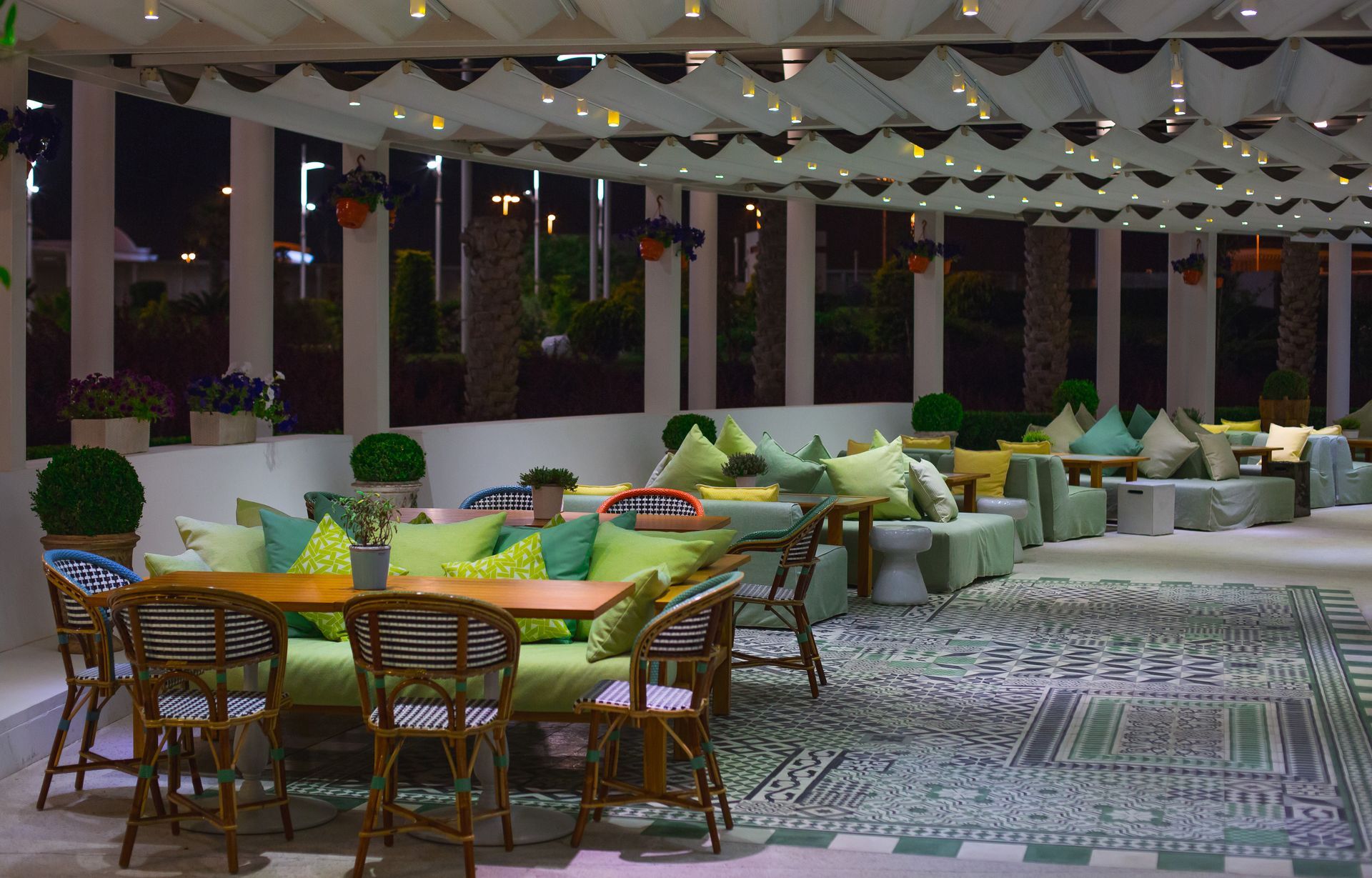 Outdoor restaurant seating at night, green cushions, wooden tables and chairs, decorative rug and ceiling.
