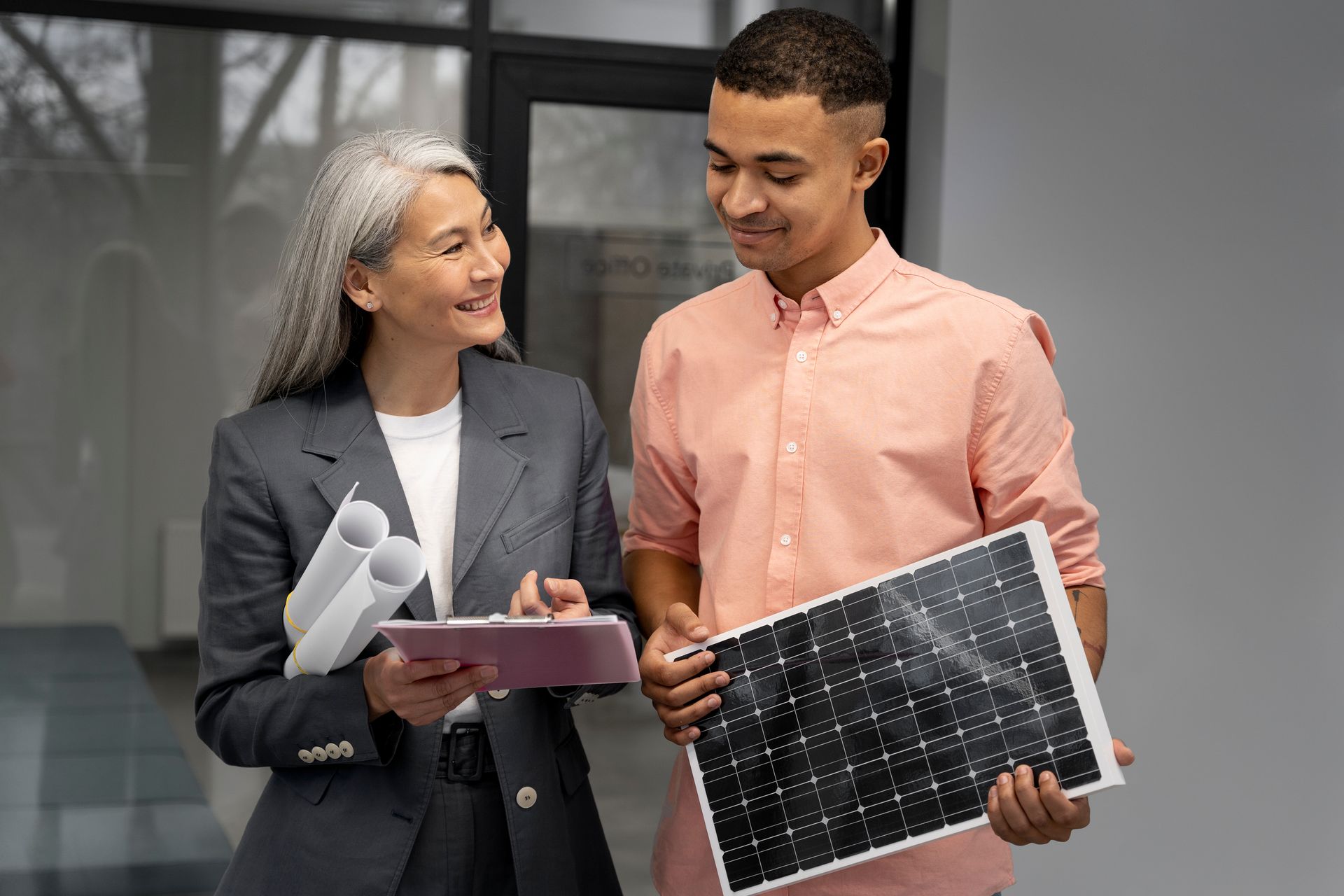 Woman in a suit and young man with a solar panel discussing plans indoors.