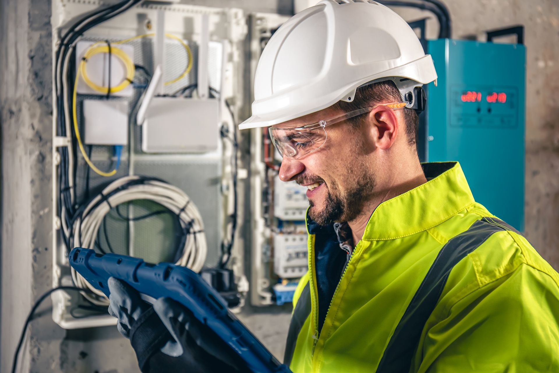 Electrician in a hard hat and safety vest, smiling while working on a circuit box.