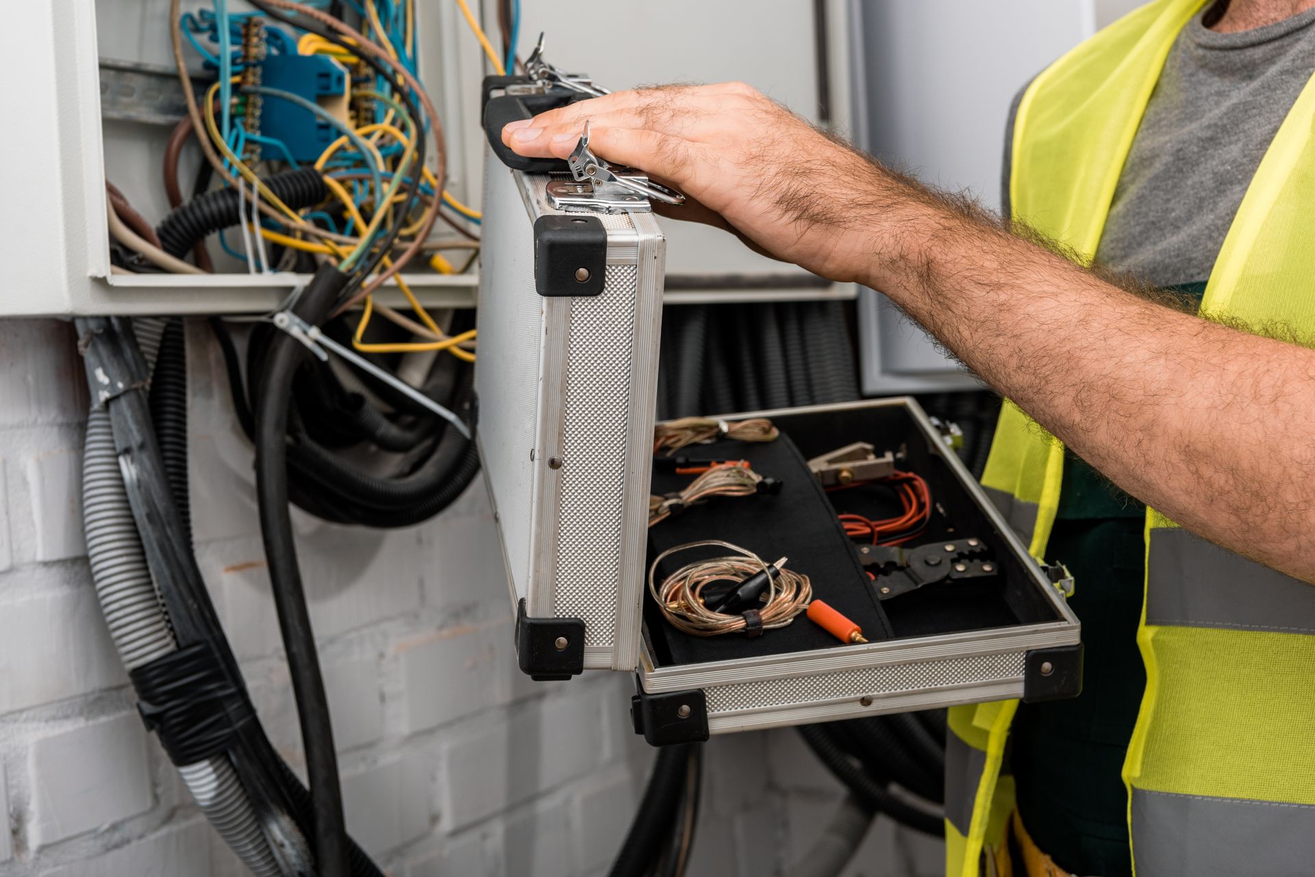 Electrician opening a metal tool case next to an open electrical wall panel with exposed wiring.
