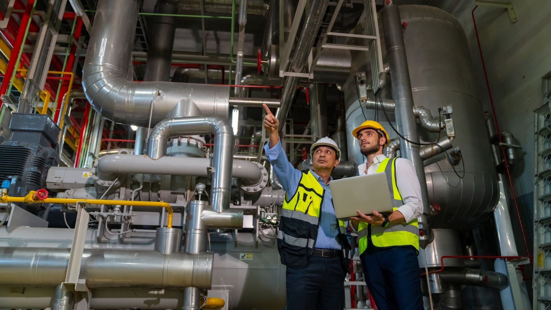 Two engineers in hard hats and vests inspecting machinery in a factory. One points, the other holds a laptop.