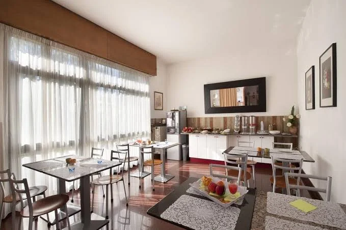 Luminosa sala da pranzo con ampie finestre, tavolo in vetro, sedie e una credenza con libri e oggetti decorativi.