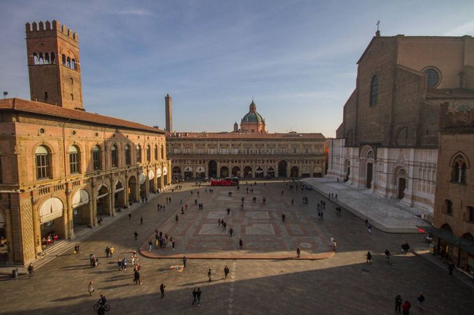 Piazza storica con edifici ad arco e una torre, persone sparse nell'ampia piazza.