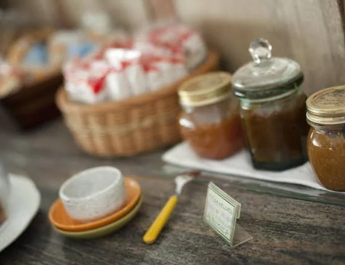 Marmellate assortite in vasetti di vetro accanto a tazze, piatti e un cesto di vimini su un tavolo di legno rustico.