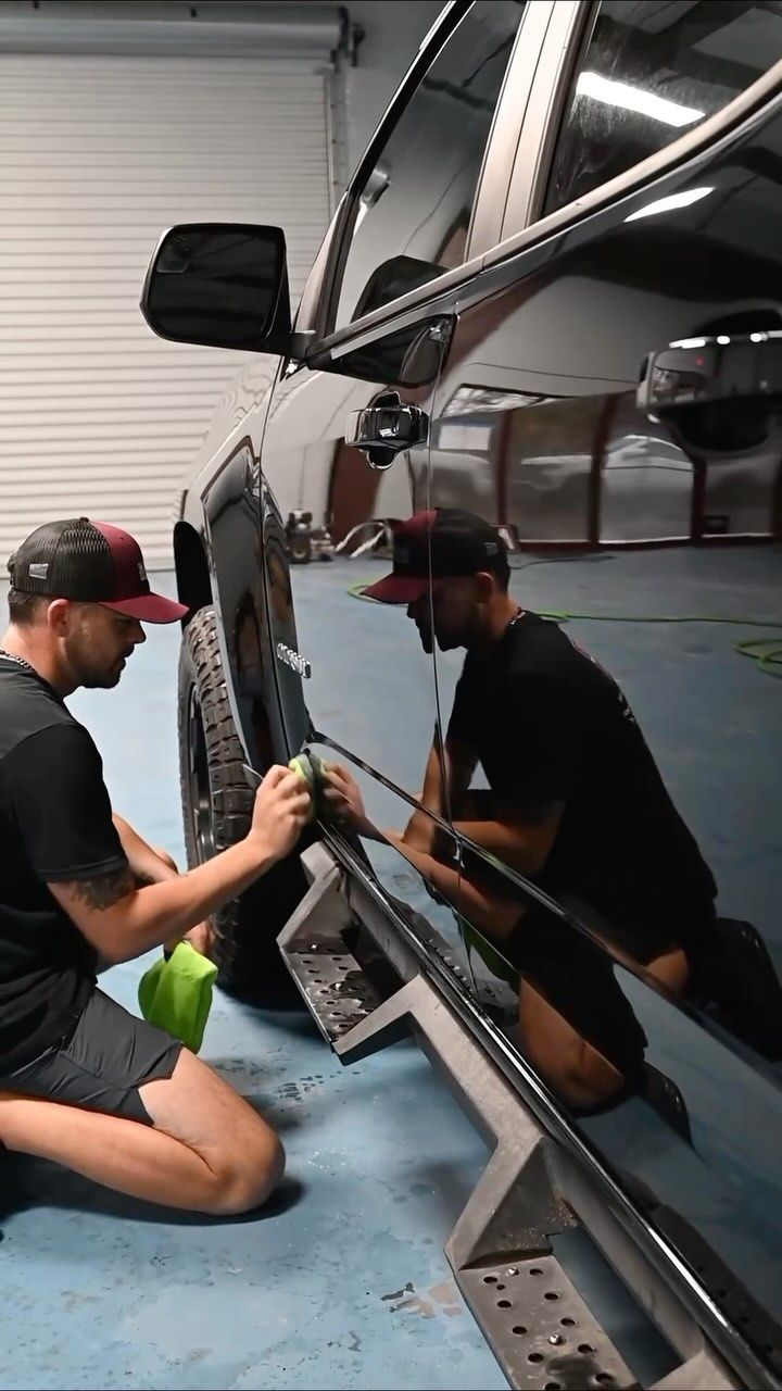 Man polishing black truck's side panel in a garage. He wears a cap, shorts, and a black shirt.