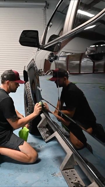 Two mechanics kneel beside a black car, polishing the side panel in a garage.