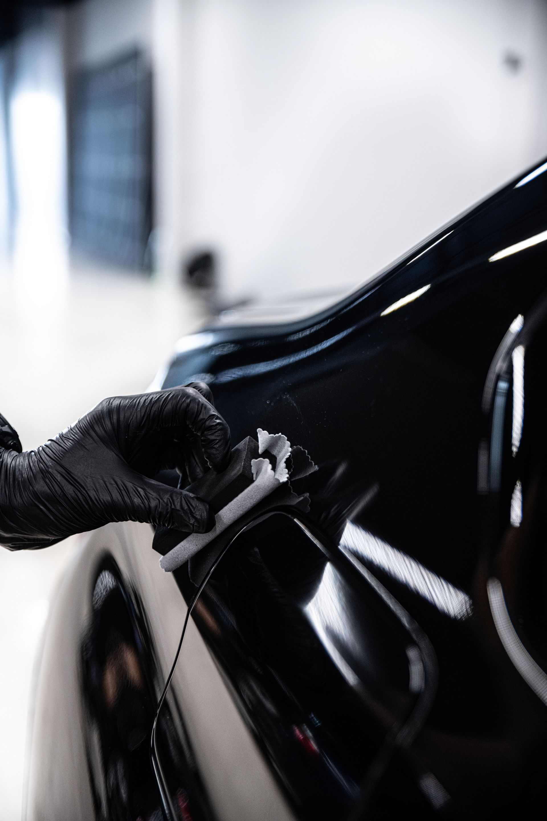 Black-gloved hand applying ceramic coating to a shiny black car in a clean, white-walled garage.