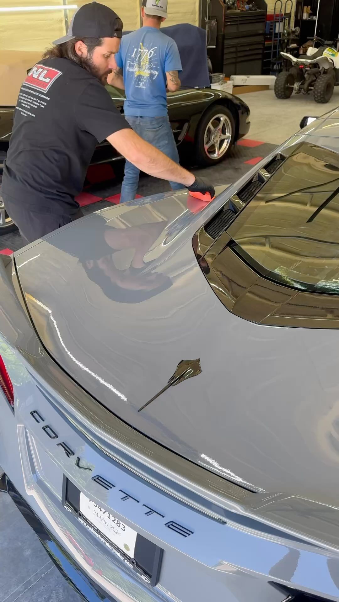 Man applying product to a gray Corvette's hood, another man in the background, shop setting.