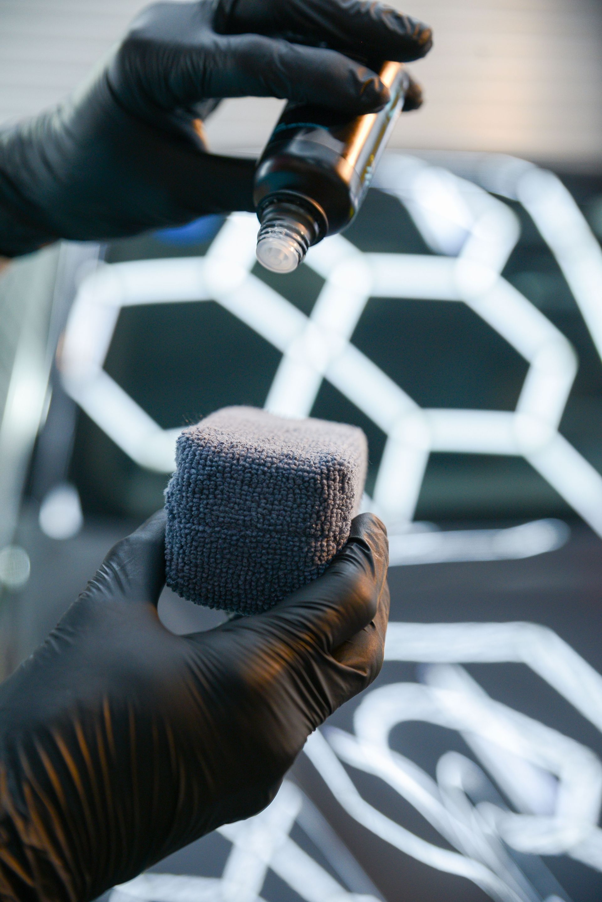 Person in black gloves applying a liquid from a bottle onto a gray applicator pad, with a car in the background.
