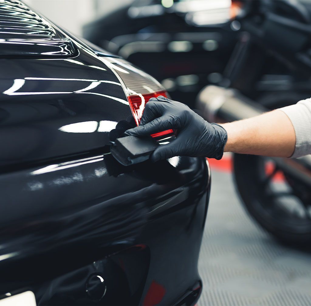 Person in black gloves applying a detailing product to a black car's taillight, motorcycle in background.