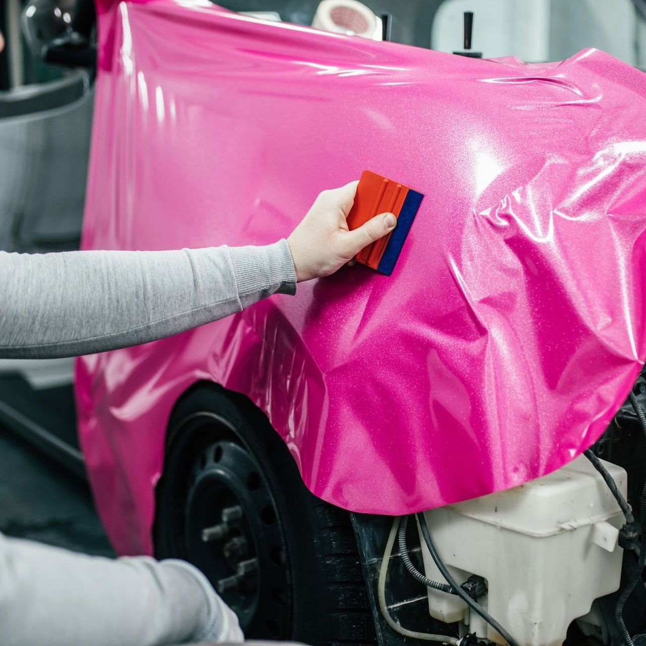 A person in a black shirt and cap polishes a black car's side panel in a garage.