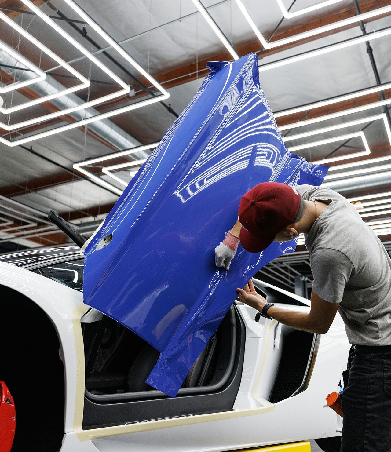 Person wrapping a blue car door on a white car inside a shop, under bright fluorescent lights.