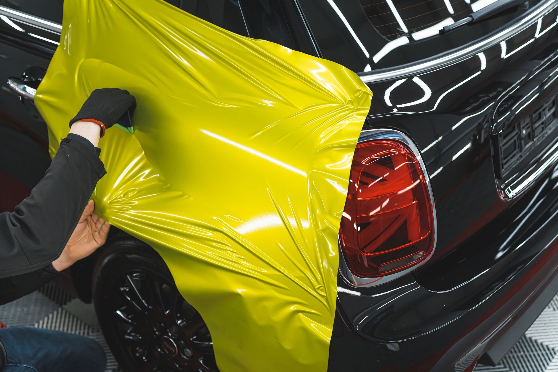 Person applying yellow vinyl wrap to a black car's rear, covering the body panel.