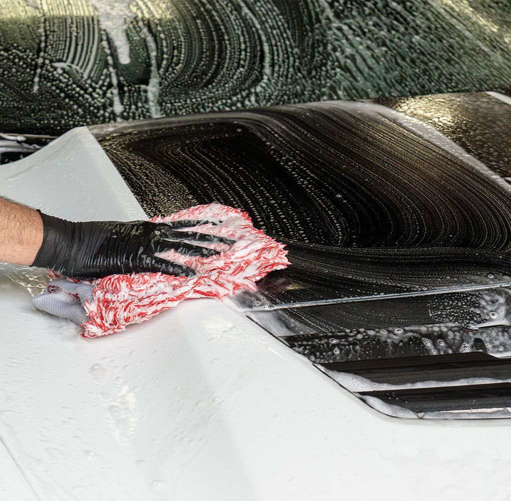 A person wearing black gloves washes a white car with a red and white rag, covered in soap suds.