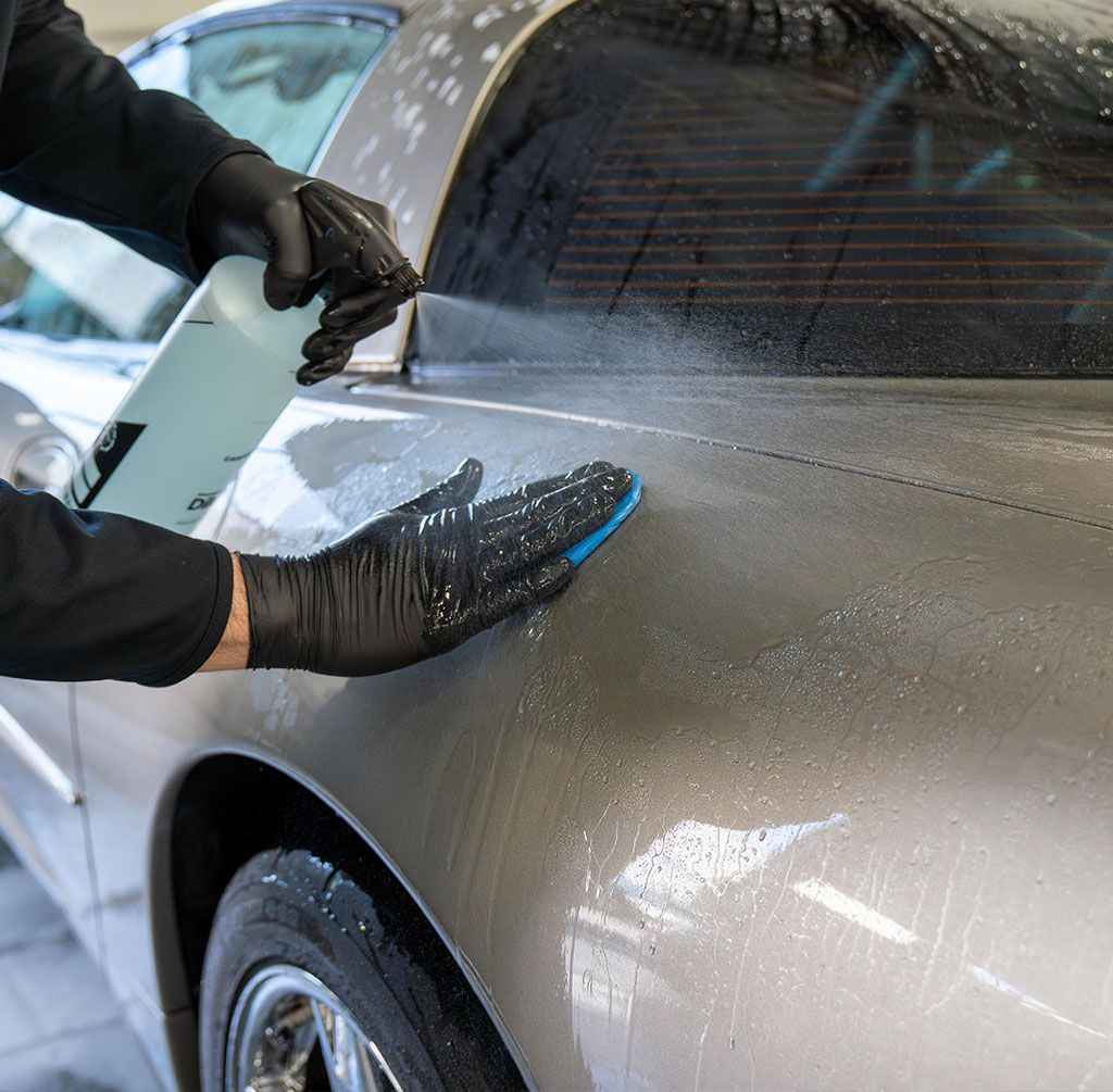 Person washing silver car, spraying solution and using sponge, wearing black gloves.