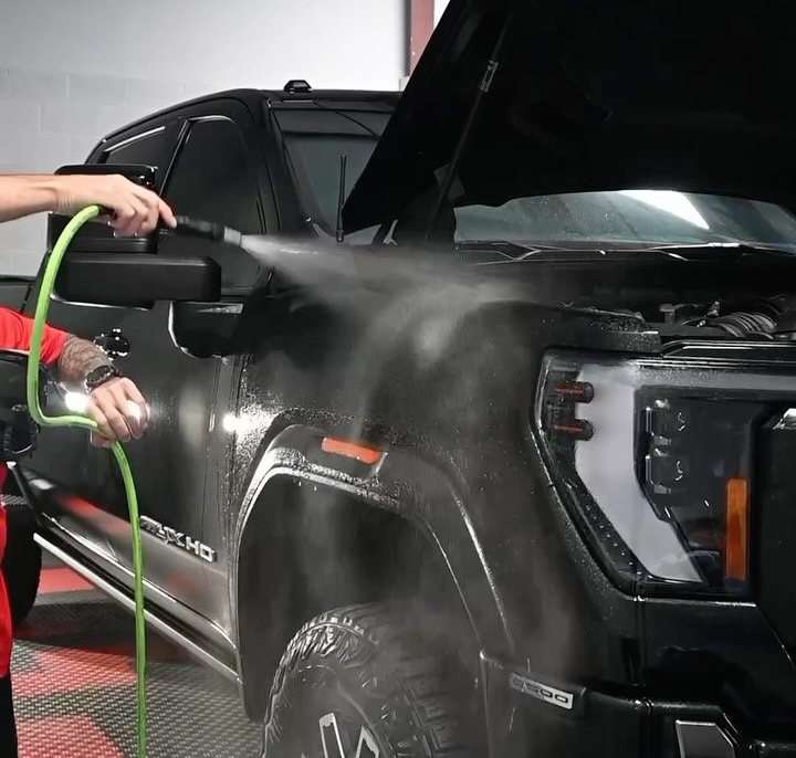 Person spraying water on a black truck with the hood open in a garage.