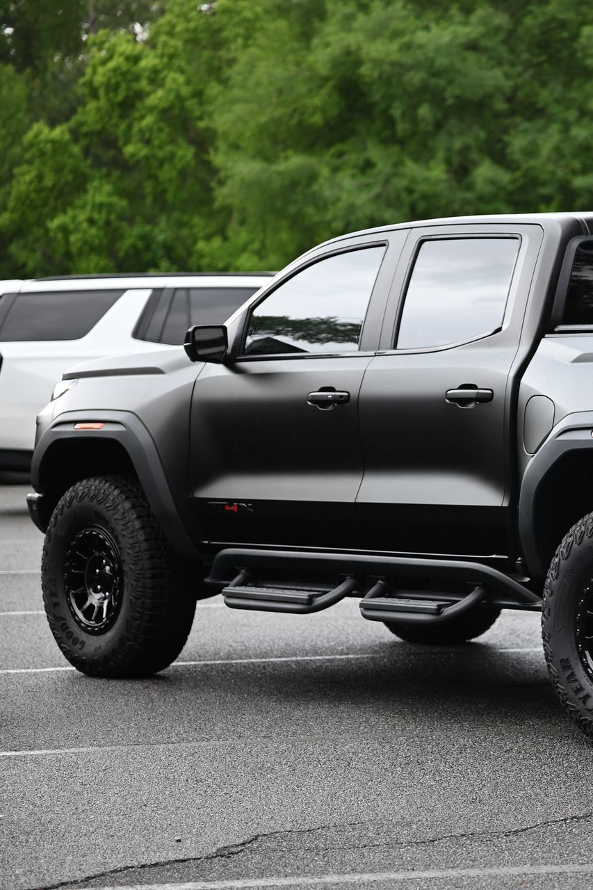 Black pickup truck parked on wet pavement with a white SUV behind it and green trees in the background