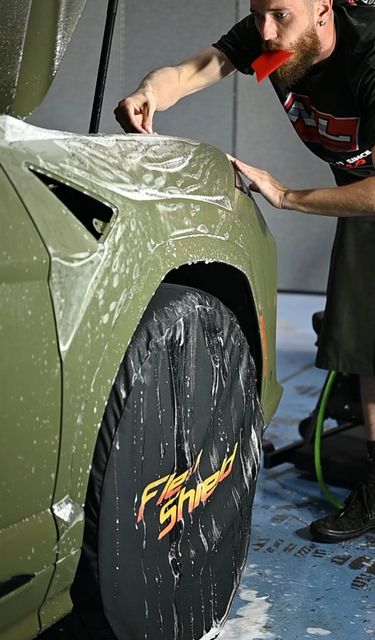 Person polishing a green car hood at an auto detail shop, with a black wheel and wet floor visible.