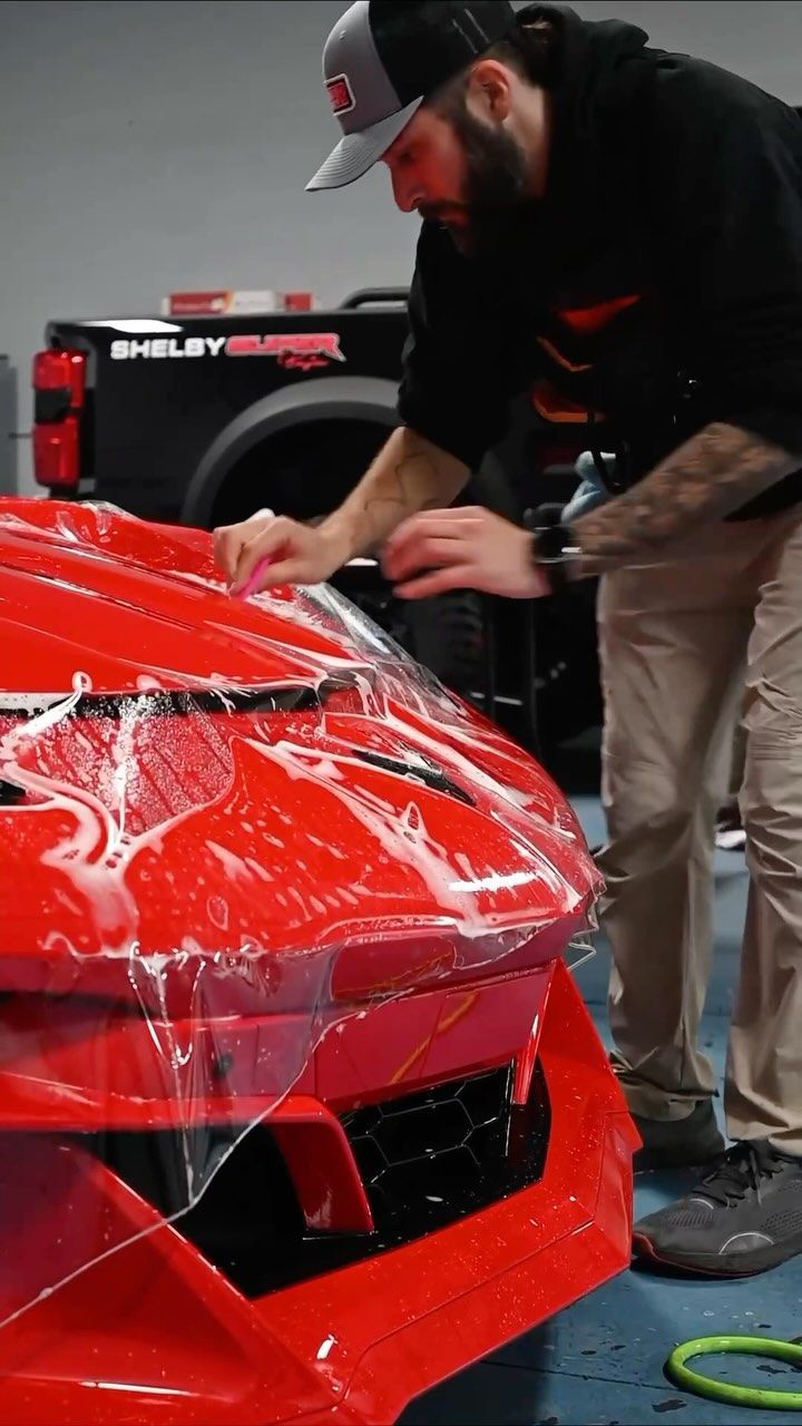 Man applying protective film to a red car hood in a shop.