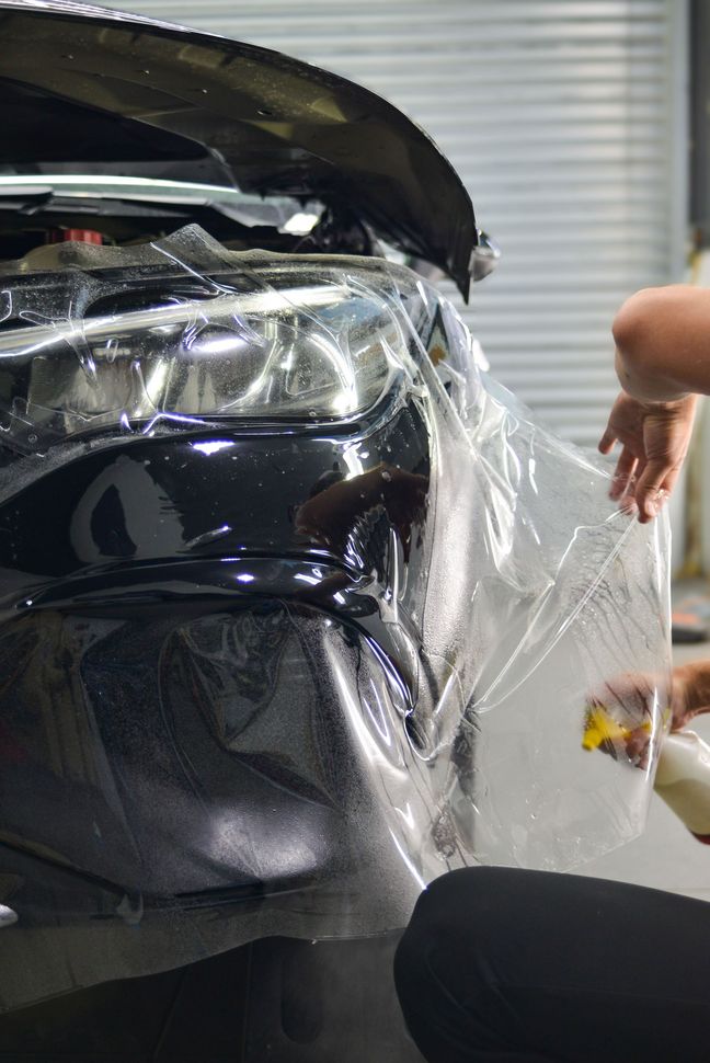 Person applying clear film to a black car's bumper in a garage.