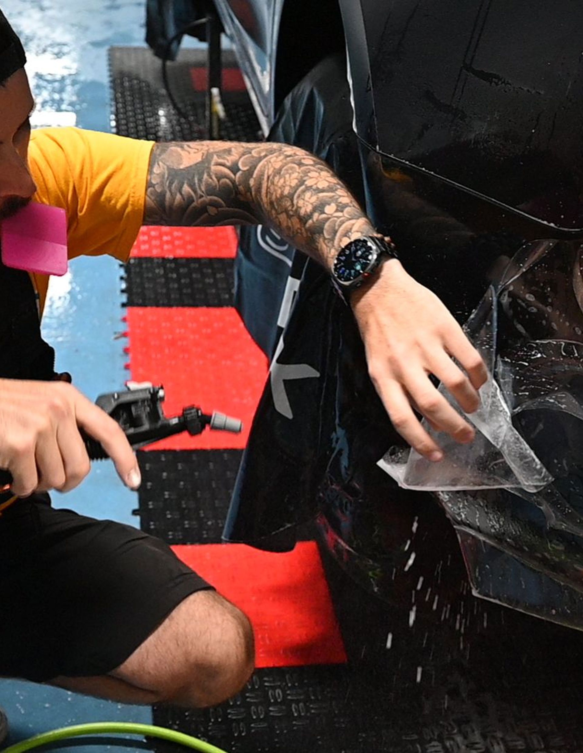 Man applying film to a car bumper with tools in a garage.