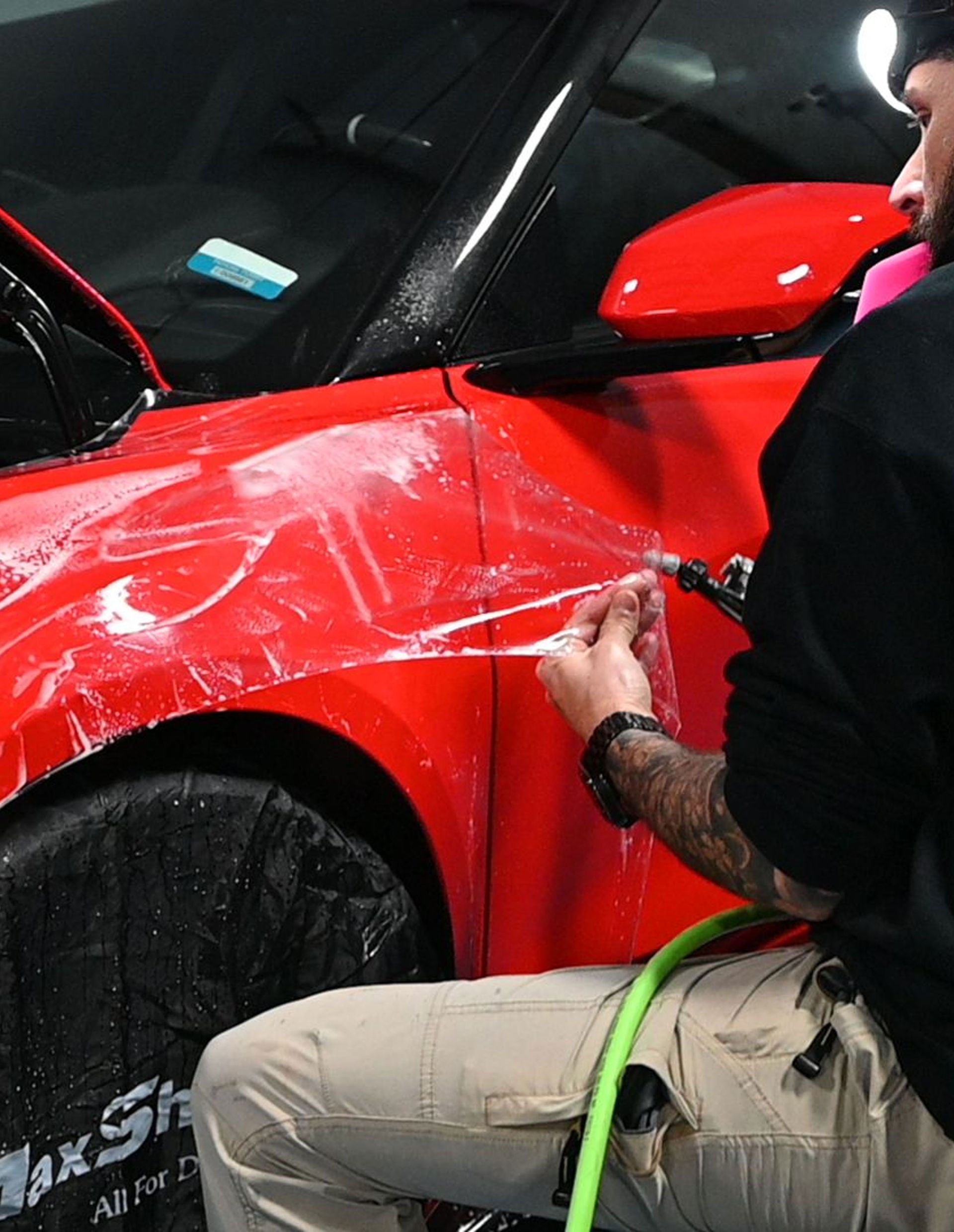 Person applying film to red car. Using tools, with green hose.
