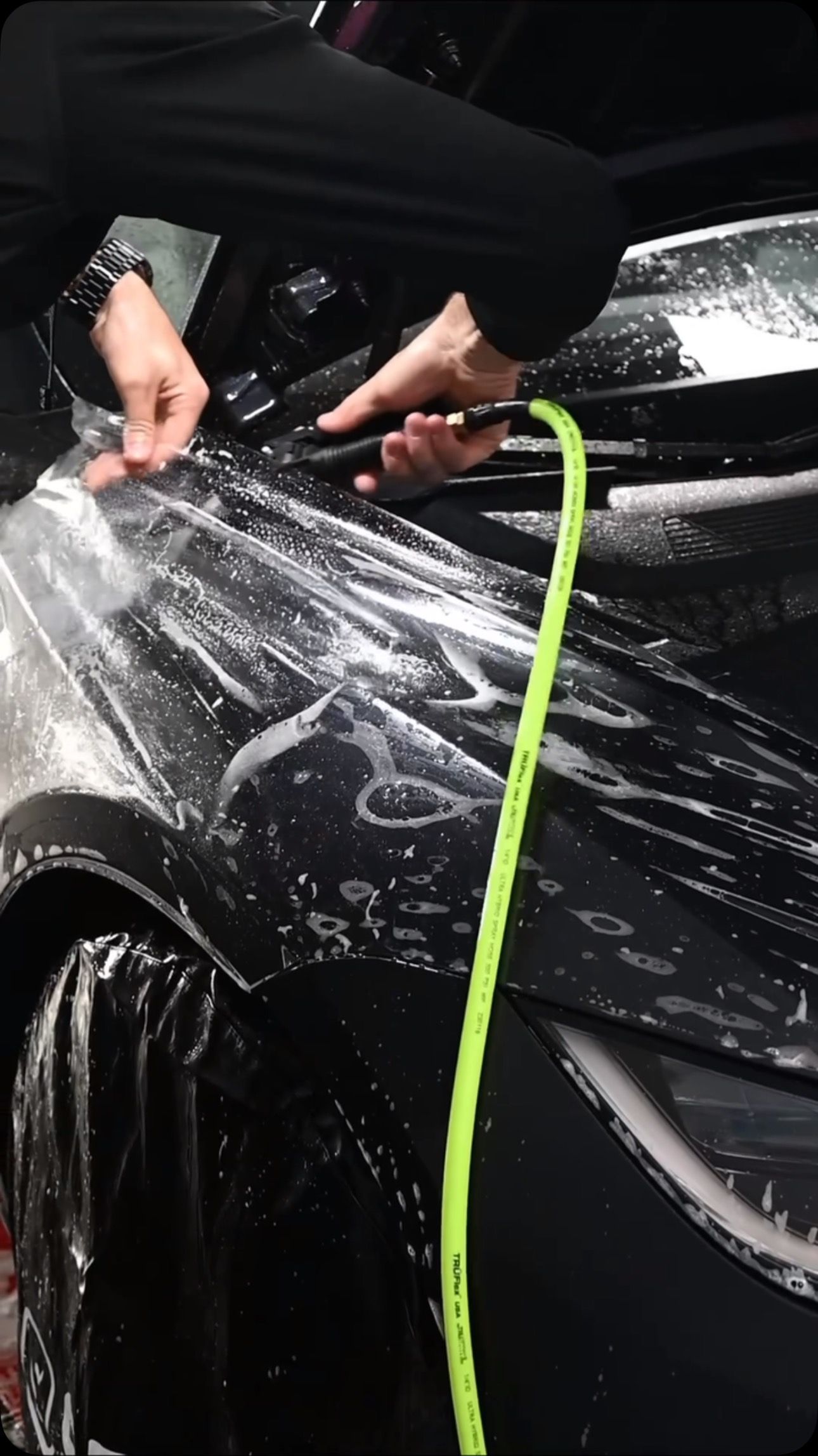 Person applying clear protective film to a black car's headlight, using tools and water spray.