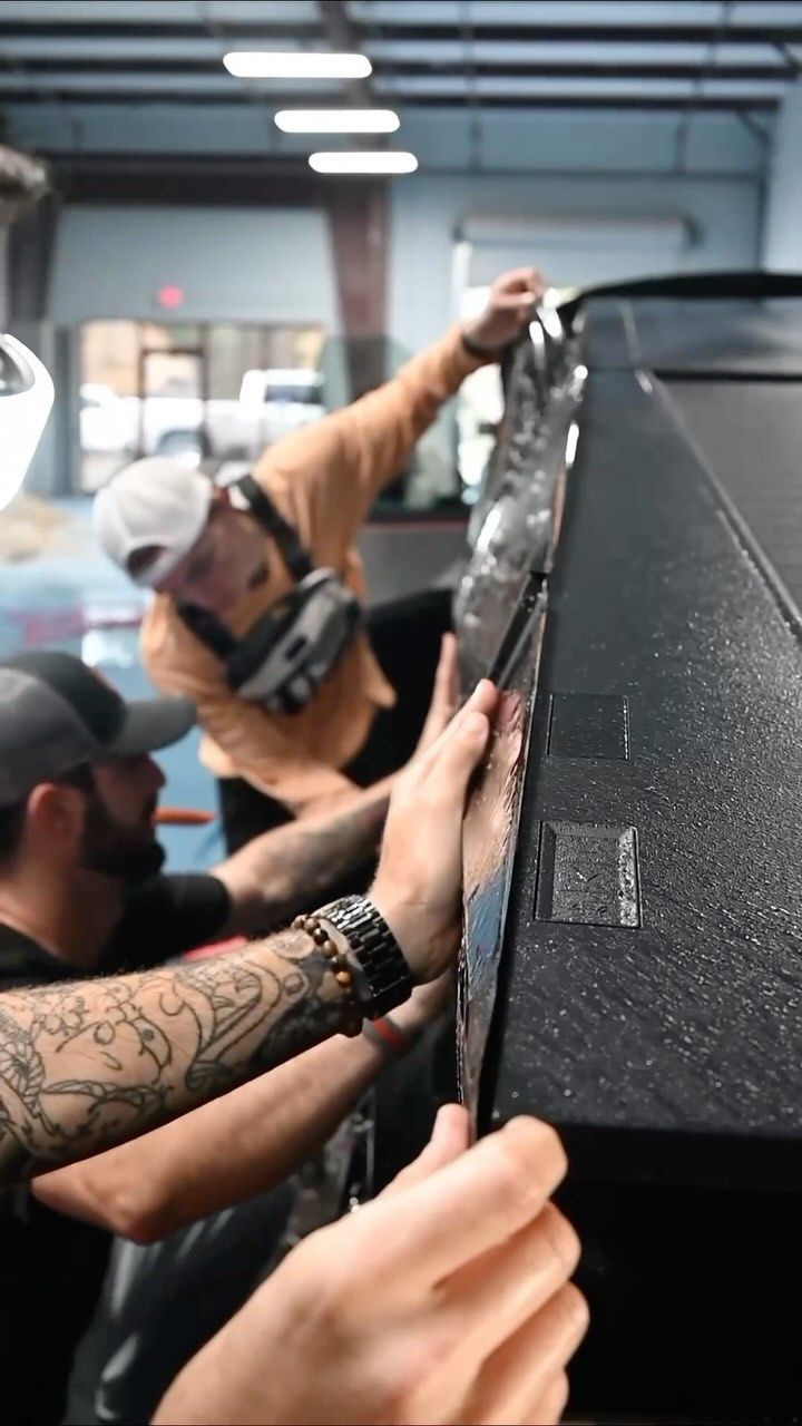 Two people applying film to the side of a black vehicle in a garage.