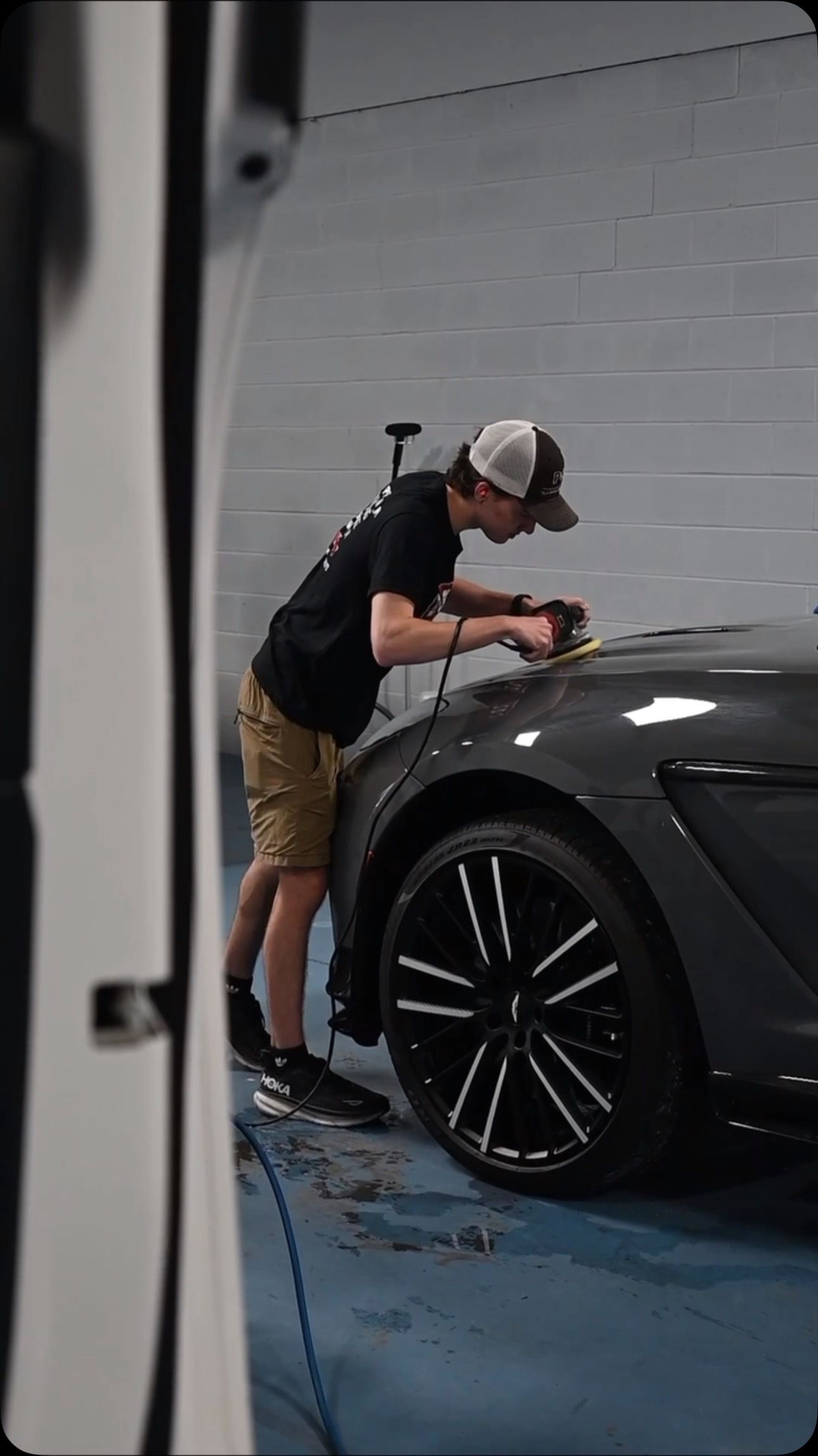 Person working on a gray car in a garage, using a polishing tool. They are wearing a hat, shorts, and a dark shirt.