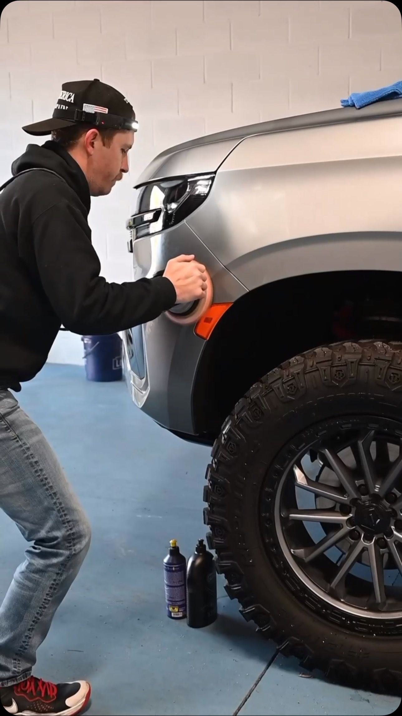 Man polishing a silver car's fender with a buffer. The car has a black tire and rim. Bottles are on the ground.