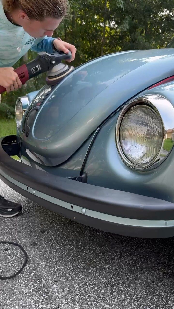 Person using a power buffer on the blue hood of a vintage VW Beetle outdoors.