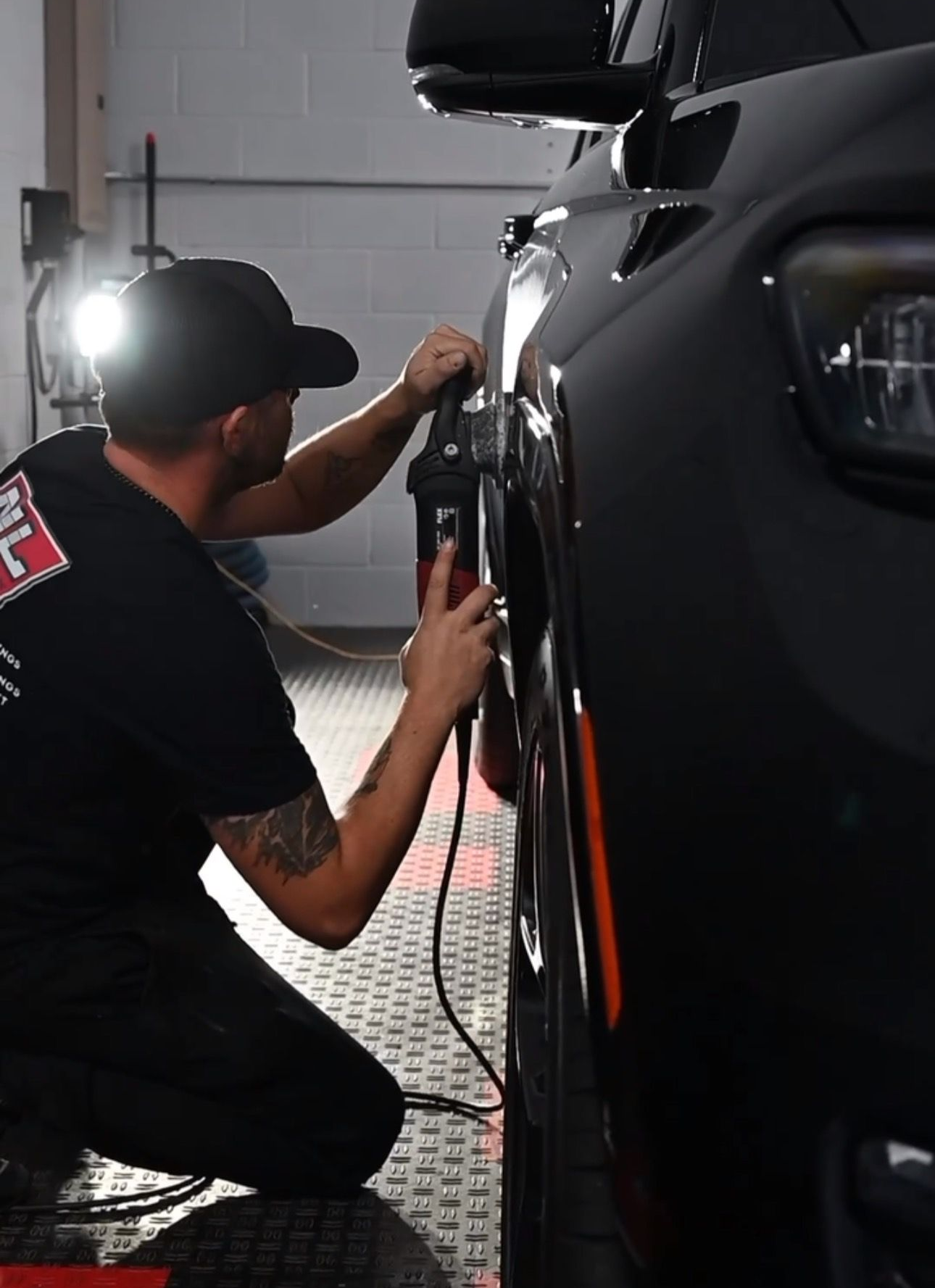 A person in a black shirt and cap polishes a black car's side panel in a garage.