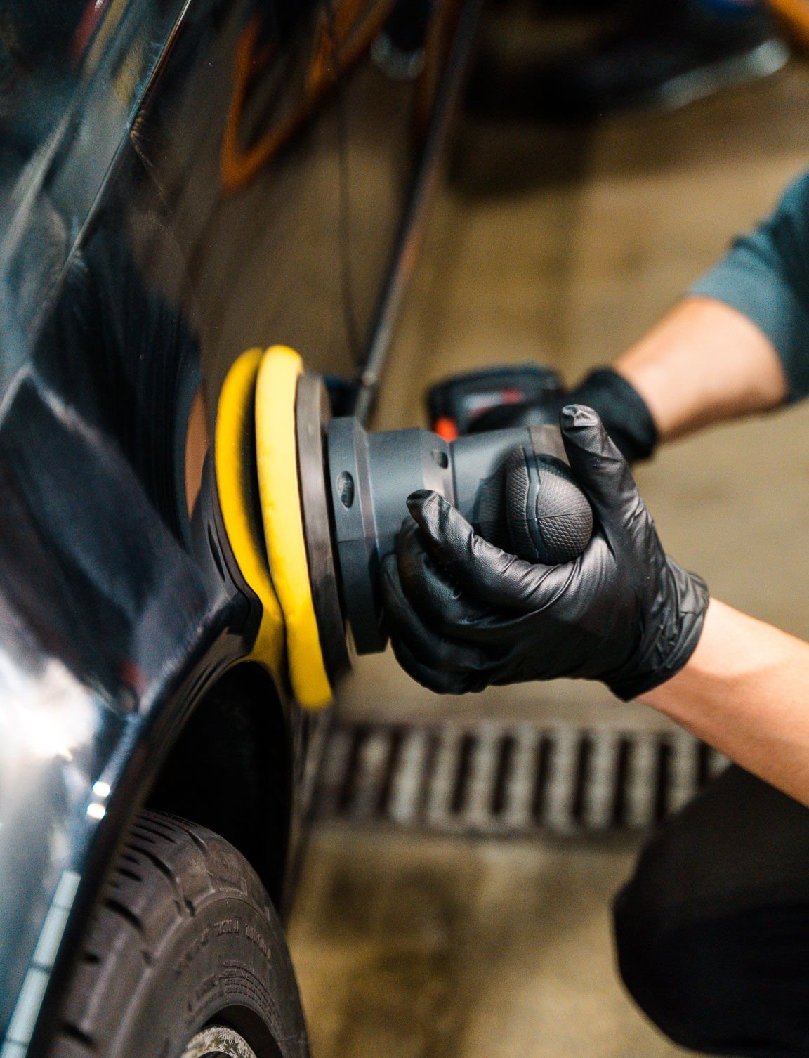 A person wearing black gloves polishes a black car with a yellow buffer in a garage.