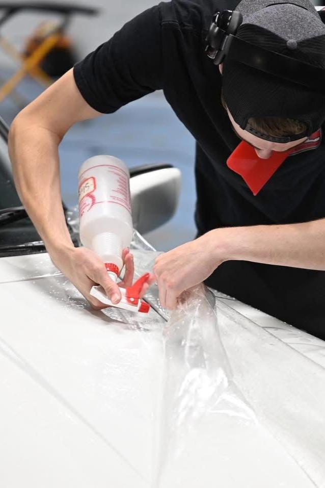 Person carefully applying red tape to a white surface with a clear tool, wearing a black shirt and cap.